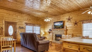 Interior de una cabaña de troncos con chimenea, muebles de madera y un televisor montado encima.
