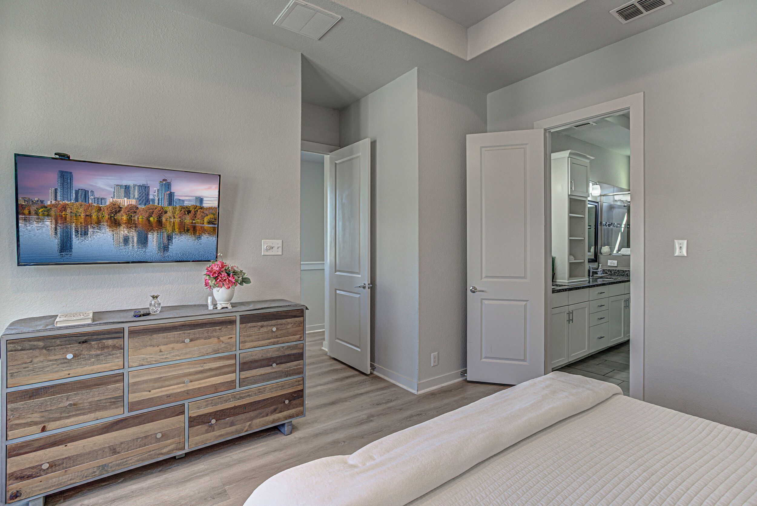 Une chambre à coucher avec un tableau représentant un paysage urbain, une commode en bois, un lit et un coin cuisine visible à travers une porte ouverte.