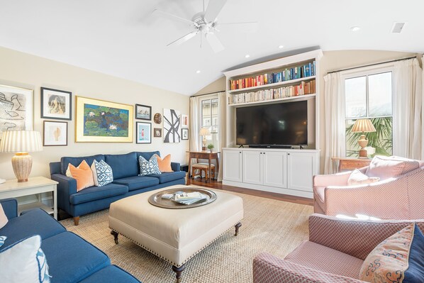 A living room with a blue sofa, a beige ottoman, a pink armchair, and a bookshelf filled with books.