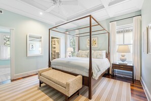 A four-poster bed with a wooden frame, a bench, a bedside table with a lamp, and a window with curtains.