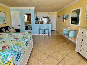 A compact hotel room with a bed, a desk, a chair, and a kitchenette.
