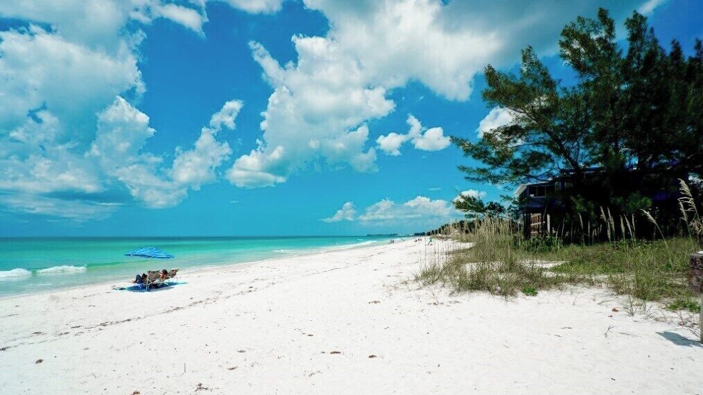 A beach with white sand, clear turquoise water, and a few people. There is a building with a porch on the right.