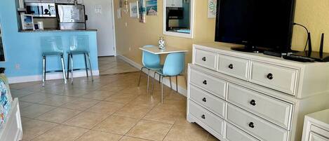 A compact living space with a kitchenette, a dining area with blue chairs, a white dresser, and a flat-screen TV.