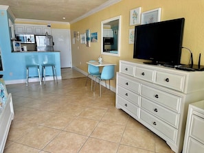 A compact living space with a kitchenette, a dining area with blue chairs, a white dresser, and a flat-screen TV.
