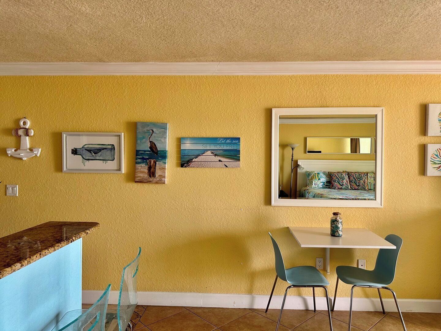 Un comedor con una mesa blanca y dos sillas color turquesa, una pared amarilla y tres cuadros enmarcados.