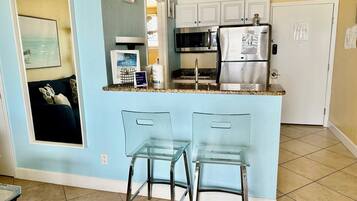 Cozinha compacta com geladeira, micro-ondas e pia. Há dois banquetas de bar de vidro e uma mesa pequena. As paredes são pintadas de azul claro, e há uma porta que dá para outro cômodo.
