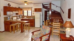 A compact kitchen with wooden cabinets, a microwave, and a refrigerator. There is a dining area with a wooden table and chairs, and a staircase leading to an upper level. - Pueblo del Mar 4☙Monkeys & Birds will make your mornings special❧ (Tamarindo)
