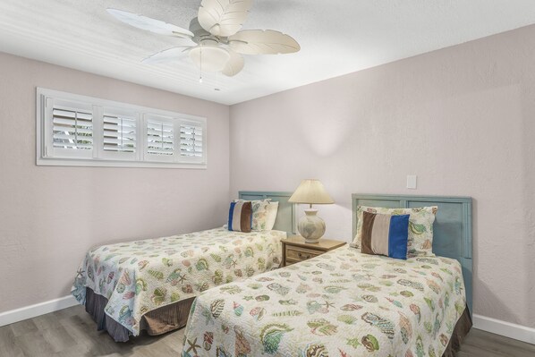 Une chambre avec deux lits, un ventilateur de plafond, une fenêtre avec des stores et une table de chevet avec une lampe.