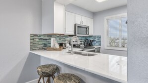 A modern kitchen with white cabinets, a stainless steel microwave, and a tiled backsplash. There are two bar stools and a window with blinds.