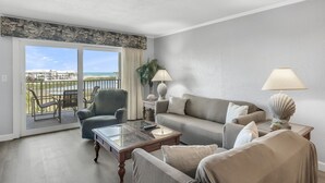 A living room with a sofa, armchairs, a coffee table, and two lamps. A balcony with a view of the ocean and buildings is visible through the glass doors.