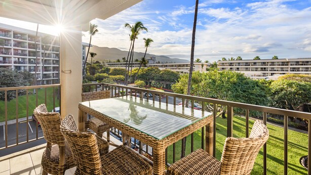 Un balcón con mobiliario de mimbre, una mesa de vidrio y vistas a un paisaje urbano con edificios y palmeras.