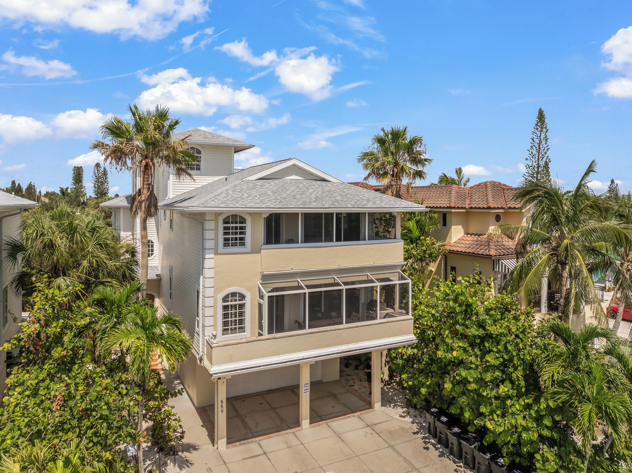 A two-story house with a covered patio, surrounded by palm trees and other greenery.