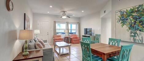 A living room with a sofa, coffee table, and chairs, a dining area with a table and chairs, a TV, and a ceiling fan.