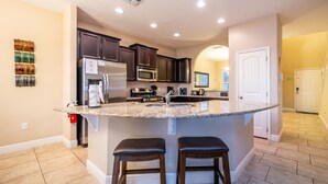 A modern kitchen with dark cabinets, a large island with stools, stainless steel appliances, and a tiled floor. - Solterra Resort Beautiful Pool Home, Spa, Lake View & Resort Amenities (Davenport)