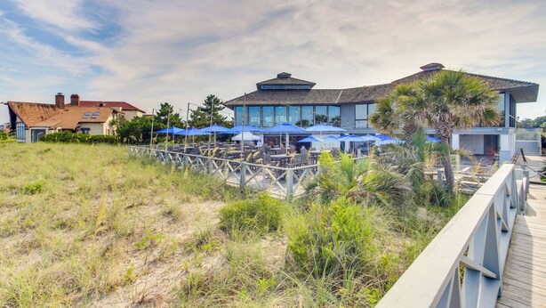 A beachfront property with a large house, outdoor seating area with umbrellas, and a wooden deck.