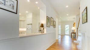 A bright hallway with wooden flooring, white walls, and a kitchen area visible through a glass door.