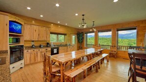 A spacious kitchen with wooden cabinets, a long dining table, and benches. There are large windows offering a view of the outdoors. - Knockin On Heavens Door: Incredible Views, Theater (Gatlinburg)