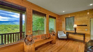 A cabin with a wooden interior, large windows with a view of green hills, a bench, a white chair, and a table with a lamp. - Knockin On Heavens Door: Incredible Views, Theater (Gatlinburg)