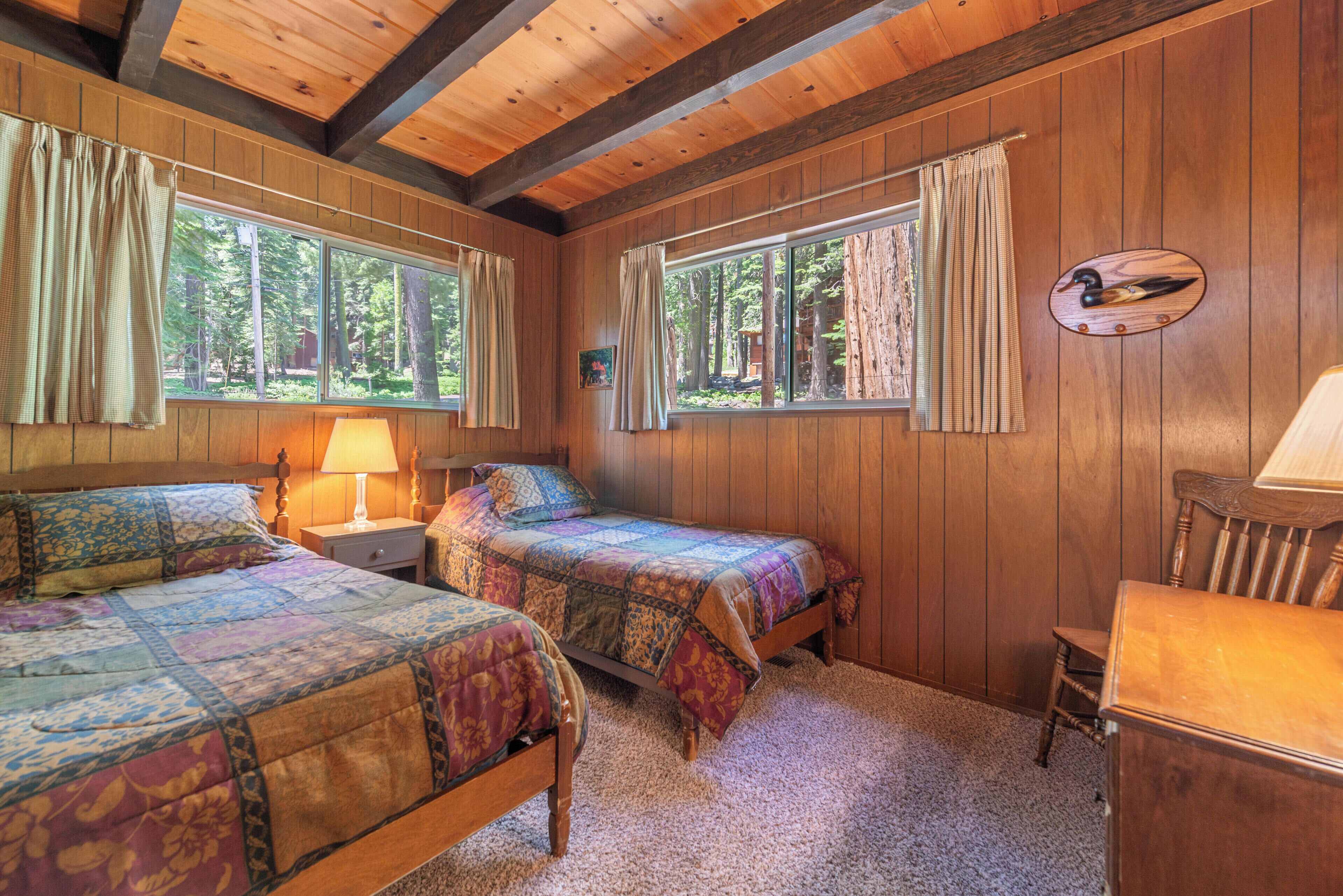 Un dormitorio de cabaña con dos camas, cabeceras de madera, una mesita de noche, una lámpara y una ventana con cortinas.