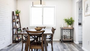 Et spiseområde med et træbord og stole, et vindue med persienner, en stige og en hylde med planter og køkkenudstyr.