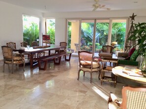 A spacious room with a large dining table, several chairs, and a sofa. There are large windows providing a view of greenery outside.