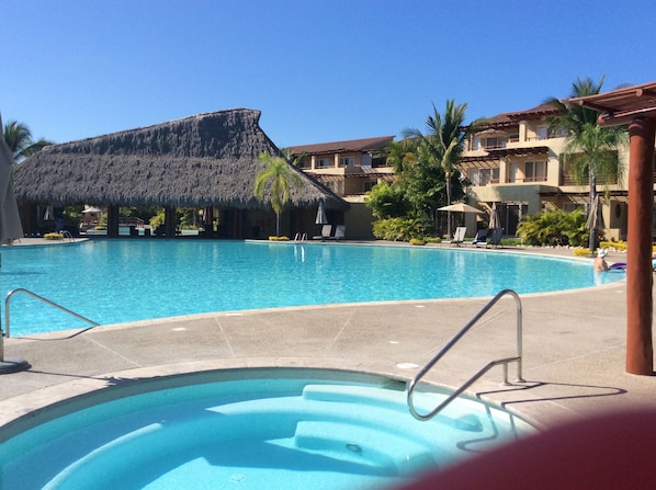 A large swimming pool with a thatched-roof structure in the background, surrounded by palm trees and lounge chairs.