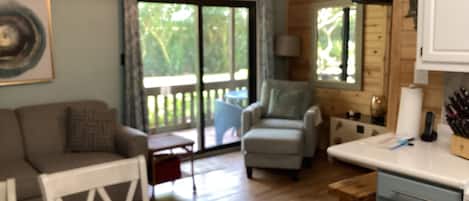 Une pièce avec un plafond en bois, un ventilateur de plafond, une table blanche, un fauteuil gris, un banc en bois et un coin cuisine équipé d’un évier et de placards.