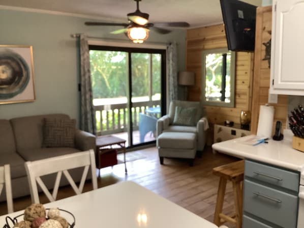 A room with a wooden ceiling, a ceiling fan, a white table, a grey armchair, a wooden bench, and a kitchen area with a sink and cabinets.