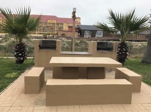 Een terras met een betonnen tafel en banken, omringd door palmbomen en een gebouw met een rood dak op de achtergrond.