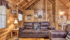 A cozy cabin interior with a wooden ceiling, a leather sofa, a wooden table, and a dining area.