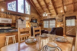 Cozy cabin-style interior with a high ceiling and wooden beams. Features a fully equipped kitchen, dining area, and comfortable seating space with a TV.