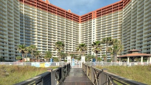 A high-rise hotel with multiple balconies, palm trees, and a wooden walkway leading to the entrance. - 6th Floor Condo with Amazing Views and Free Activities This Summer! (Panama City Beach)