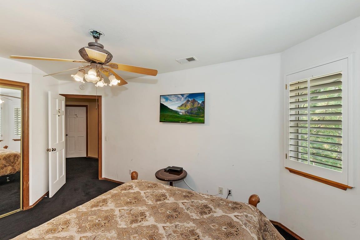 Un dormitorio con cama, ventilador de techo, un cuadro, ventana con persianas y puerta que da a otra habitación.