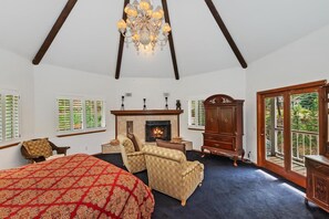 A bedroom with a large bed, two armchairs, a fireplace, and a view through a glass door.