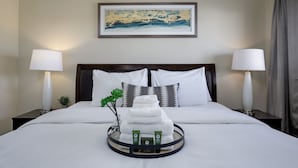 Una cama bien hecha con sábanas blancas, dos lámparas de noche, una obra de arte enmarcada sobre la cama y una bandeja con artículos de aseo en la mesita de noche.