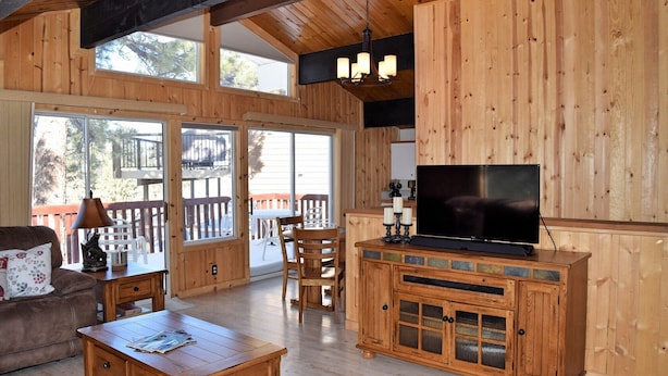 A cozy cabin interior with wooden walls, a large TV on a wooden cabinet, a dining area with a table and chairs, and a sofa.