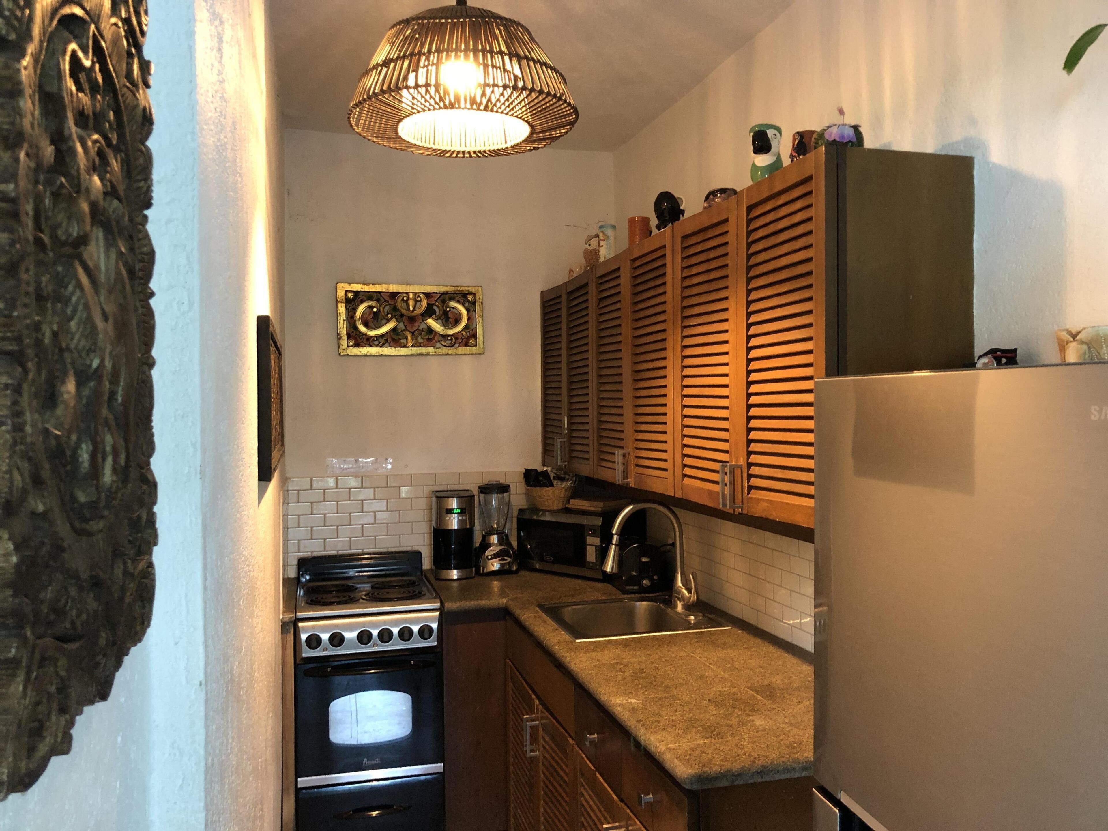 Een compacte keuken met een kookplaat, oven, spoelbak en een koelkast. Boven de kookplaat hangt een decoratief wandobject en boven de spoelbak houten keukenkastjes.