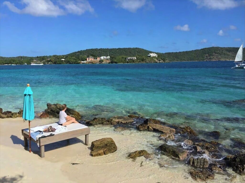 A beach with a bench and a blue umbrella, clear turquoise water, and a sailboat in the distance.