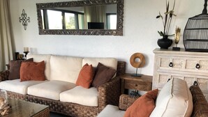 Uma sala de estar com um sofá bege, almofadas marrons, uma mesa de centro de vime, uma mesinha lateral com um prato decorativo e um armário branco com gavetas.