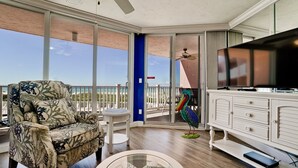 A balcony with a view of the beach, a sofa, a table, a colorful bird sculpture, and a TV.