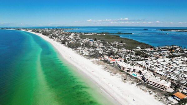 An aerial view of a coastal town with a long sandy beach, clear turquoise waters, and a mix of residential and commercial buildings.