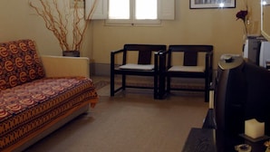 Una sala de estar con un sofá, dos sillones, una ventana con contraventanas, un jarrón con ramas secas y un cuadro en la pared.