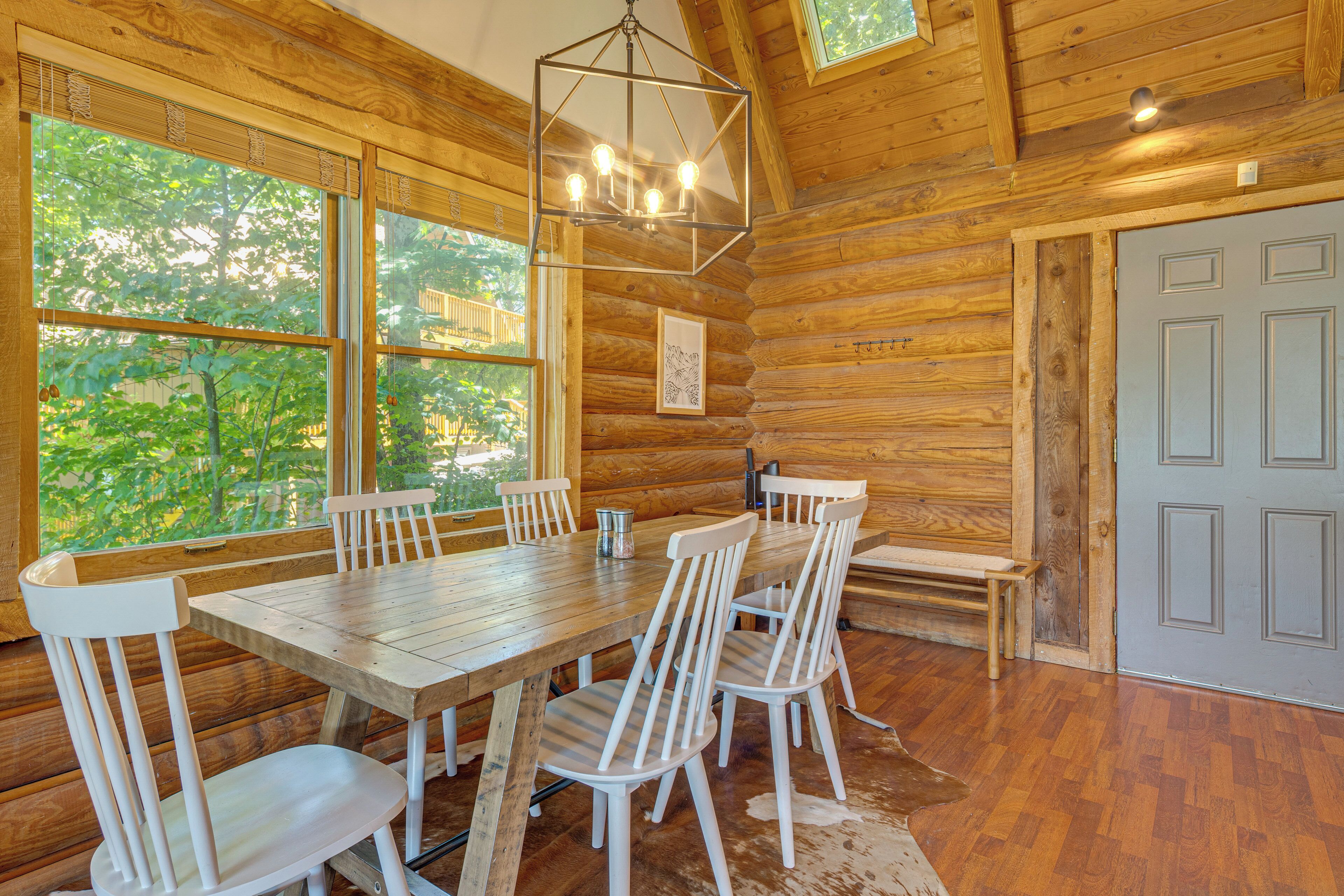 A wooden cabin interior with a dining table, chairs, a bench, and large windows.