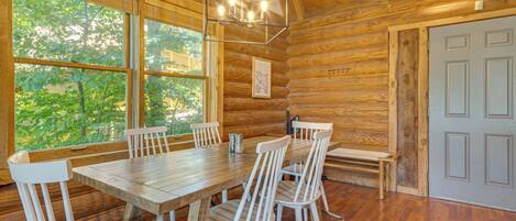 A wooden cabin interior with a dining table, chairs, a bench, and large windows.