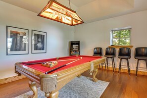 Une table de billard recouverte d’un feutre rouge, un queue de billard et un jeu de billes. Trois chaises noires sont placées à droite de la table.