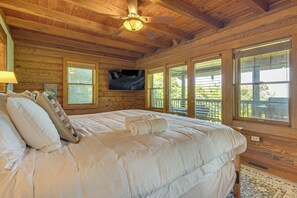 Camera da letto in una baita di legno, con un letto, una ventola a soffitto, una TV e ampie finestre.