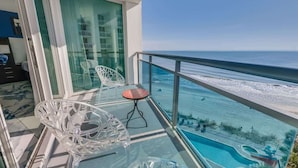 A balcony with a glass table and chairs, overlooking a pool and beach.