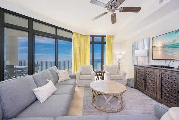 A modern living room with a gray sofa, two armchairs, a wooden coffee table, and a TV on a wooden cabinet. Large windows offer a view of the ocean.