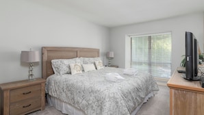 A bedroom with a large bed, two bedside tables, a TV, and a window with blinds.
