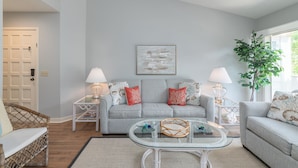 A living room with a grey sofa, two side tables with lamps, a glass coffee table, a wicker chair, and a large plant.
